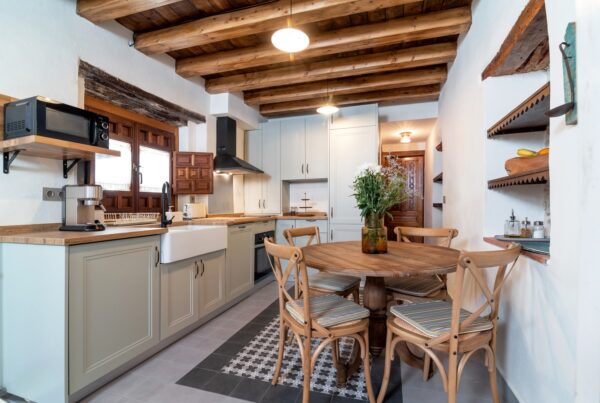Cozy farmhouse-style kitchen featuring a wood-beam ceiling, shaker cabinets, open shelving, and a round table with chairs.
