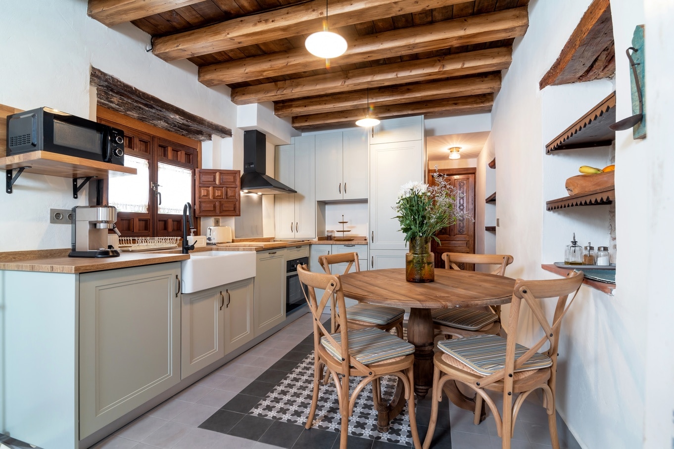 Cozy farmhouse-style kitchen featuring a wood-beam ceiling, shaker cabinets, open shelving, and a round table with chairs.