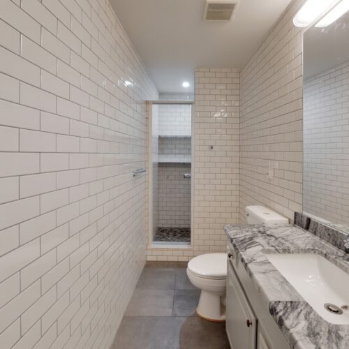home improvement contractors White-tiled bathroom featuring a gray marble sink, mirrored wall, toilet, and open shower niche beyond.