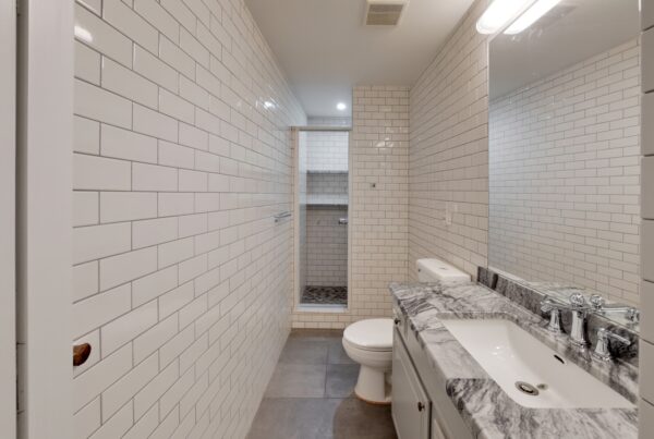 White-tiled bathroom featuring a gray marble sink, mirrored wall, toilet, and open shower niche beyond.