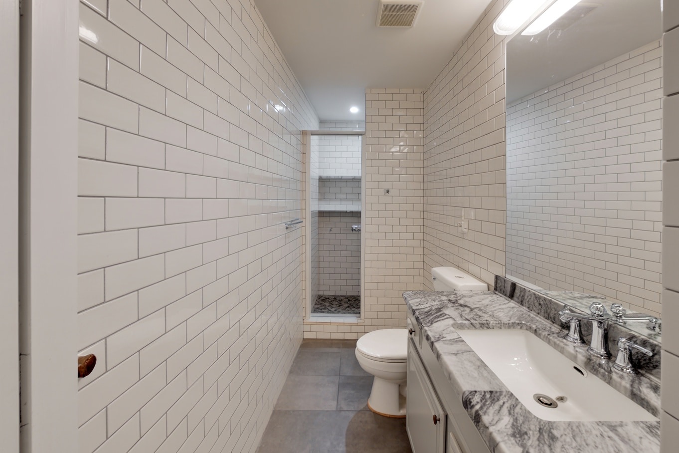 White-tiled bathroom featuring a gray marble sink, mirrored wall, toilet, and open shower niche beyond.