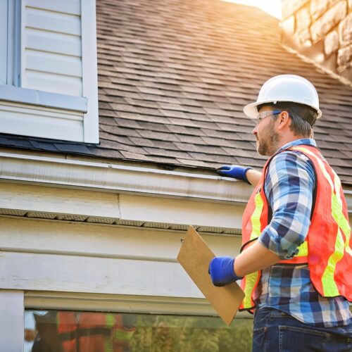 home improvement contractors A person in a white hard hat stands beside a house roof while holding a clipboard near a gutter in sunlight.