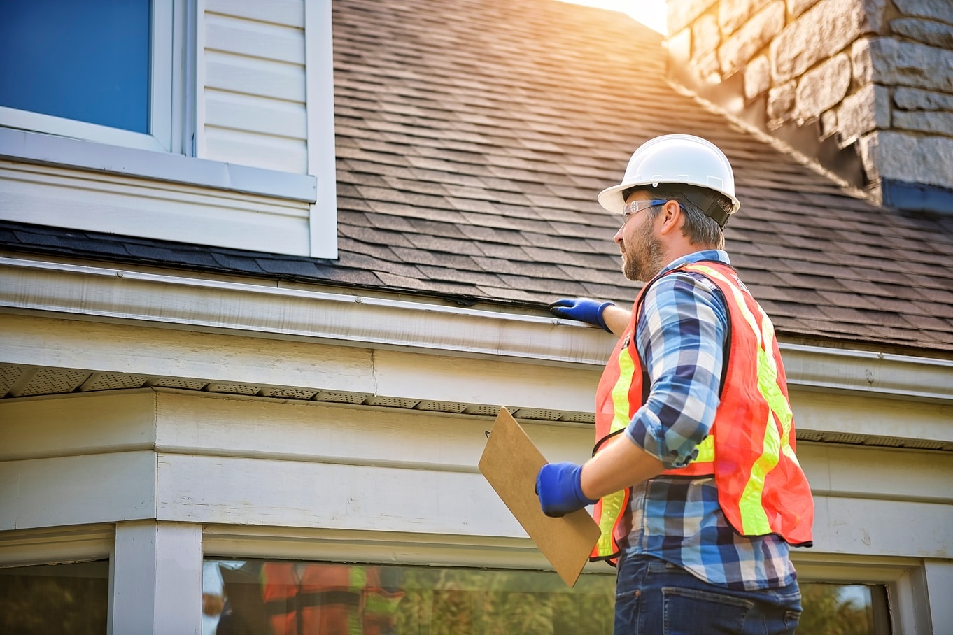 A person in a white hard hat stands beside a house roof while holding a clipboard near a gutter in sunlight.