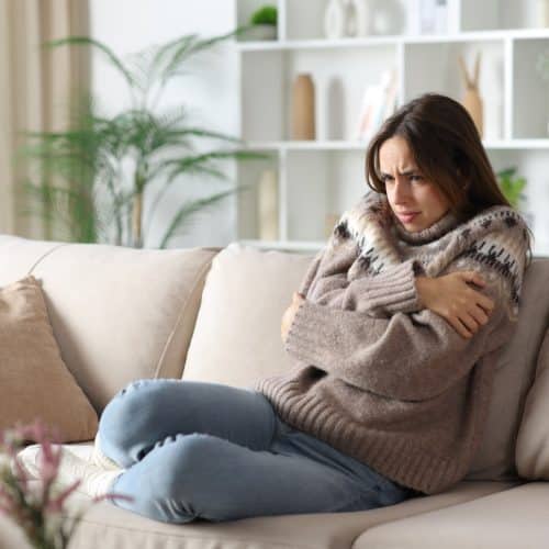 helloproject A woman wearing a thick sweater sitting on a beige sofa. She has her arms crossed because she is cold.