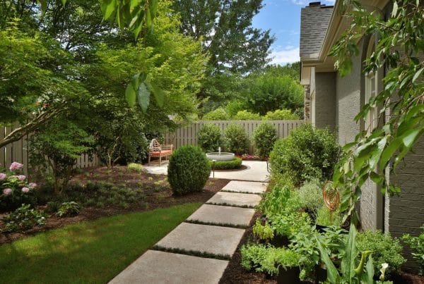 A backyard with a stone path leading to a patio that is shrouded with green trees. There is a fence around the yard.