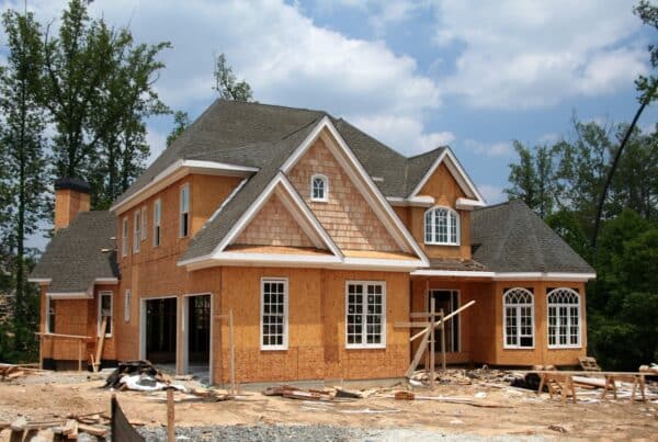 Two-story house under construction with exposed wood sheathing, a shingled roof, and building materials scattered across the yard.