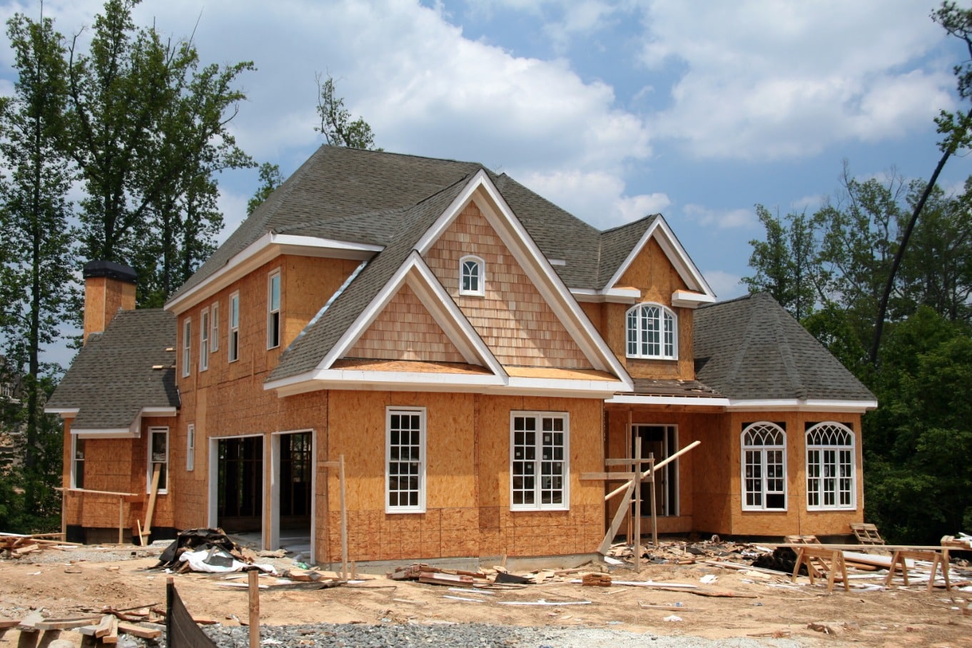Two-story house under construction with exposed wood sheathing, a shingled roof, and building materials scattered across the yard.
