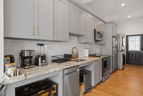 A modern galley kitchen with grey cabinets, gold hardware, stainless-steel appliances, wooden floors, and white walls.