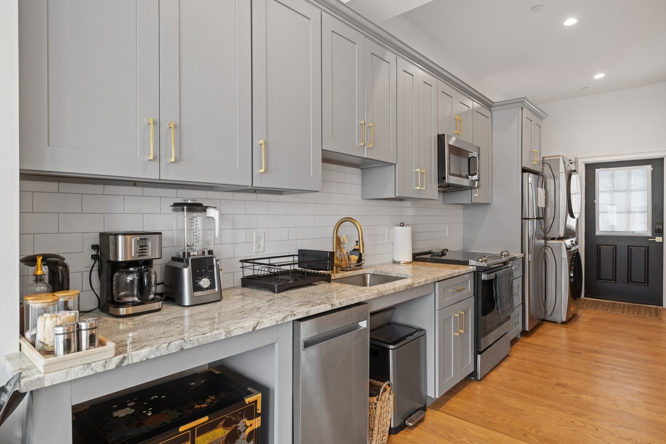 A modern galley kitchen with grey cabinets, gold hardware, stainless-steel appliances, wooden floors, and white walls.