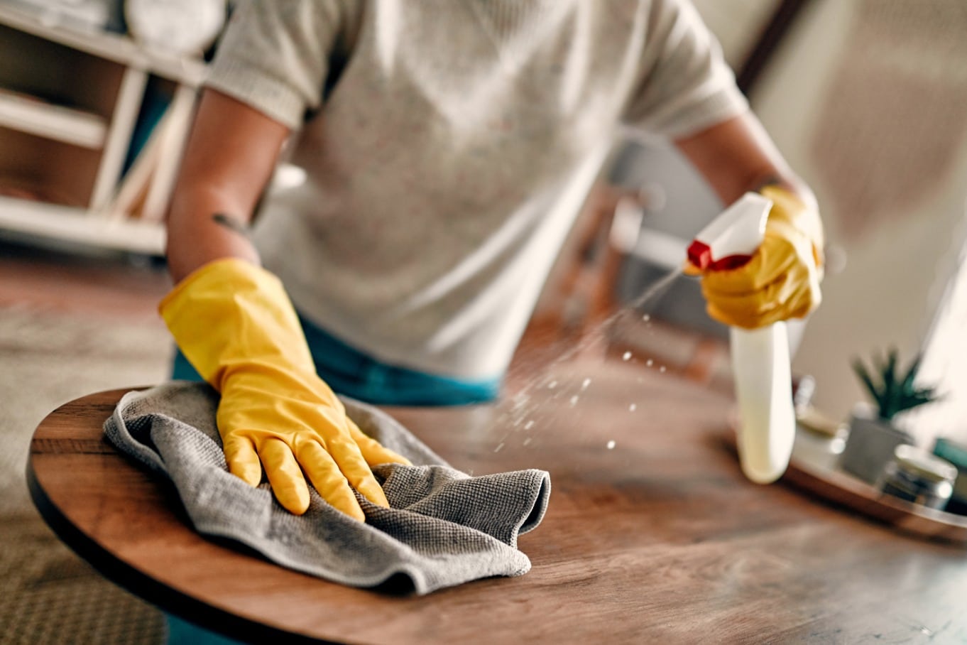 A woman is wearing yellow gloves as she uses a spray bottle and cleaning rag to clean a tabletop.